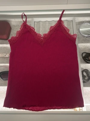 Mörk Rosa ribbad linnetopp från Amisu - Säljer en Mörk Rosa ribbad linnetopp från Amisu med tunna axelband och spetsdetaljer längs v-ringningen. Toppen är ärmlös och har en mjuk, stretchig känsla som gör den superbekväm. Perfekt för sommardagar eller att styla lager på lager.