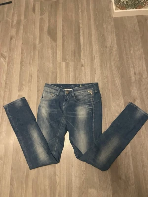 Replay blå jeans med slitningar - Säljer ett par snygga blå jeans från Replay med coola slitningar och tvättade detaljer. Klassisk femficksmodell med raka ben och normal passform. Jeansen har metallknapp och dragkedja framtill samt Replay-logga på bakfickan. Perfekt för dig som gillar streetstil.