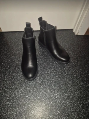 Svarta chelsea boots i skinn - Säljer ett par svarta chelsea boots med rund tå och platt sula. Skorna har elastiska panel på ena sidan och dragkedja på andra för enkel påtagning. Tillverkade i slätt skinn med diskret söm och låg klack. Perfekta för dig som gillar stilrena och klassiska boots.