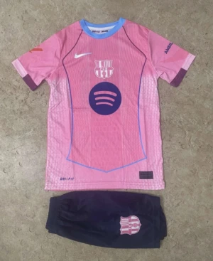 FC Barcelona fotbollströja Raphinha - Säljer en FC Barcelona matchtröja med Raphinha 11 på ryggen, i rosa med lila detaljer och blå krage. Tröjan har klubbmärke och sponsortryck på bröstet, samt tillhörande svarta shorts med klubbmärke. Perfekt för dig som älskar fotboll och Barca.