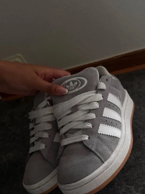 Grå Adidas sneakers med vit sula - Säljer ett par gråa Adidas sneakers med klassiska vita ränder på sidan och vit logga på plösen. Skorna har vit snörning och en gummisula i beige ton. Ovandelen är i mocka och har en rund tå. Perfekt för dig som gillar stilrena och bekväma sneakers.