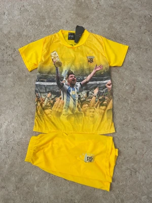 Gul Messi fotbollsset Argentina - Säljer ett gult fotbollsset med Messi och nummer 10 på ryggen. Tröjan har tryck av firande med VM-pokalen och AFA-logga, och matchande shorts med samma logga. Perfekt för dig som älskar fotboll och Argentina.