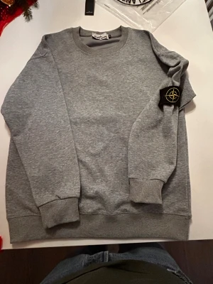 Grå sweatshirt från Stone Island - Säljer en grå sweatshirt från Stone Island med klassisk rund hals och ribbade muddar. Tröjan har den ikoniska Stone Island-patchen med gul brodering på vänster ärm, fäst med knappar. Perfekt för dig som gillar stilrena och snygga streetwear-plagg. Köpt ifrån Kauf Haus de Westens i Berlin ( som NK fast i Tyskland)