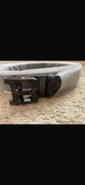 Svart bälte med mönster - Säljer en svart väst med ett stort B-format spänne framtill. Västen har ett diskret rutigt mönster med små B-detaljer och är tillverkad i ett syntetmaterial med en glansig finish. Passformen är normal och designen är stilren med fokus på detaljer.