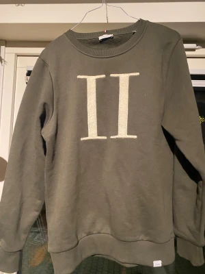 Olivgrön sweatshirt Les Deux - Olivgrön sweatshirt från Les Deux med stort vitt II-tryck i frotté på bröstet. Tröjan har rund halsringning, ribbade muddar och är långärmad. Perfekt för dig som gillar stilrena och bekväma plagg med en twist.