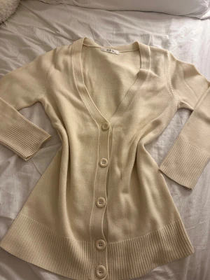 Beige kofta med v-ringning från SPB - Snygg beige kofta från SPB med djup v-ringning och stora knappar framtill. Modellen har långa ärmar med breda ribbade muddar och ribbad nederkant. Perfekt att slänga över en t-shirt eller linne för en chill look.