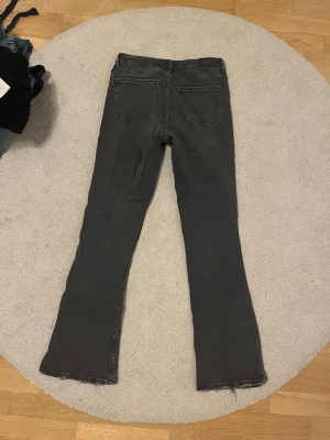 Svartgråa lågmidjade bootcut jeans från Gina Tricot - Bootcut jeans från Gina Tricot i svart tvättad denim. Modellen har låg midja, klassiska fem fickor och rå kant vid benslut. Materialet är bomull med stretch för skön passform. Snygga detaljer med knappar och dragkedja framtill. Aldrig använda! Bara skriv om du har några frågor!🩷
