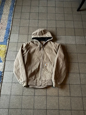 Beige sandstone washed Carhartt active jacket L - Säljer en beige Carhartt active jacket i storlek L med dragkedja och huva. Jackan har ribbade muddar, två stora fickor fram och klassisk Carhartt-logga på bröstet. Materialet är slitstark bomull och insidan är fodrad i svart. Perfekt för året runt.