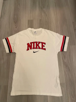 Vit Nike t-shirt med röda detaljer - Vit t-shirt från Nike med stort rött NIKE-tryck och svart swoosh på bröstet. Ärmarna har röda och svarta ränder som ger en sportig vibe. Klassisk passform och rund halsringning. Perfekt för dig som gillar streetwear och sportiga looks.
