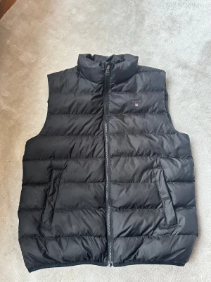 Svart dunväst från GANT - Svart dunväst från GANT med quiltad design och ståkrage. Västen har dragkedja framtill och två sidofickor. Perfekt att slänga över en hoodie för en chill och snygg look. Regular fit och diskret logga på bröstet.stlr 176
