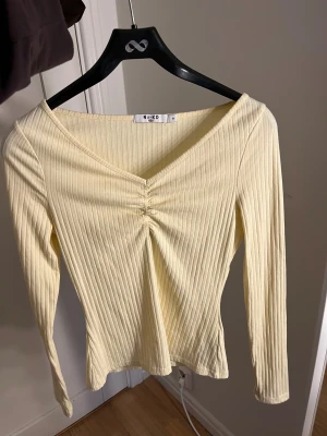 Ribbad ljusgul topp från NA-KD - Säljer en ljusgul ribbad topp från NA-KD i storlek M. Toppen har v-ringning med snygg rynkdetalj framtill och långa ärmar. Materialet är mjukt och stretchigt.