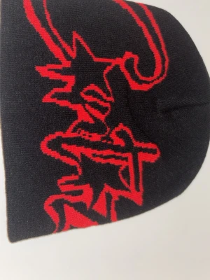 Cortez mössaa - Säljer en svart stickad mössa med ett stort rött grafiskt motiv på framsidan. Mössan är mjuk och har en klassisk form som sitter skönt på huvudet. Perfekt för dig som gillar streetstyle och vill ha något som sticker ut.