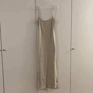Beige långklänning med smala axelband - Säljer en stilren beige långklänning med smala axelband och rundad halsringning. Klänningen har en enkel och rak siluett som ger en avslappnad men ändå elegant look. Materialet känns mjukt och följsamt mot huden, perfekt för varma dagar.