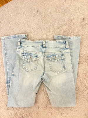 Bootcut ljusblå jeans med fickdetaljer - Säljer ett par ljusblå bootcut jeans med snygga fickdetaljer bak och klassisk femficksdesign. Jeansen har låg midja och är tillverkade i mjukt denim-material. Passar dig som gillar en avslappnad och trendig stil.