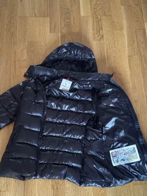 Svart Moncler maya äkta - Säljer en svart pufferjacka från Moncler med glansig finish och huva. Jackan har dragkedja framtill, två sidofickor med dragkedja och ett snyggt Moncler-märke på ärmen. Perfekt för kalla dagar och har en cool, modern look.