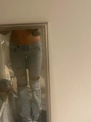 Ljusblå ripped jeans med bootcut - Säljer ett par ljusblå jeans med slitna detaljer och hål på knäna. Modellen har bootcut och normal passform, med klassiska fem fickor och knapp- samt dragkedja framtill. Jeansen är tillverkade i denim och har en cool, avslappnad vibe.