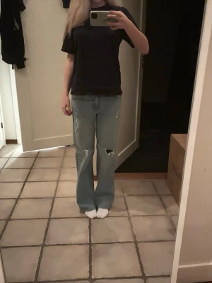 Ljusblå bootcut jeans med slitningar - Säljer ett par ljusblå jeans med bootcut passform och snygga slitningar på knäna. Jeansen har klassisk femficksdesign, låg midja och är tillverkade i mjukt denim. Perfekta för dig som gillar en avslappnad och trendig stil.