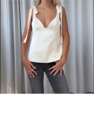 Topp  - Supersnygg vit linnetopp från Adoore i glansigt sidenmaterial. Toppen har djup v-ringning och breda axelband som knyts i rosetter. Perfekt för sommarens alla tillfällen och ger en chic och trendig look.