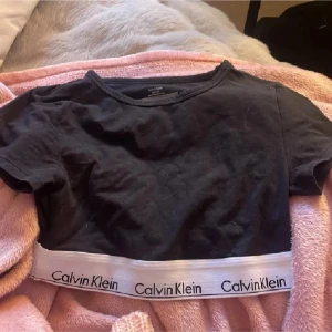 Svart croppad topp Calvin Klein - Svart croppad topp från Calvin Klein med korta ärmar och rund hals. Nedtill finns en bred vit resår med Calvin Klein-logga i svart. Toppens material är mjuk bomull och den har en tight passform som sitter snyggt mot kroppen.