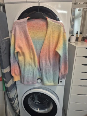 Pastellfärgad stickad kofta - Säljer en supermysig stickad kofta i härliga pastellfärger med mjuka övergångar mellan rosa, gul, blå, lila och orange. Koftan har långärmade ärmar och är öppen framtill utan knappar. Perfekt för dig som gillar färg och vill ha en chill vibe på din outfit.