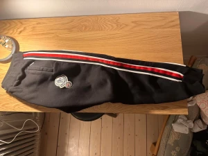 Svarta mjukisbyxor från Moncler - Svarta mjukisbyxor från Moncler med broderad logga på benet. Snygga röda och vita ränder längs sidorna, ribbade muddar vid benslut och midja samt ficka med dragkedja. Perfekta för chill eller streetwear. Materialet är mjukt och bekvämt bomull.