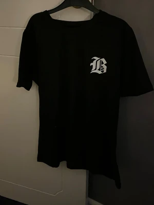 Svart t-shirt med tryck - Svart t-shirt med korta ärmar och ett vitt tryck i form av bokstaven 'B' på bröstet. Enkel och stilren design som passar perfekt till streetwear. Materialet känns som bomull och t-shirten har en normal passform.
