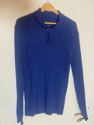 Blå ribbad tröja Massimo Dutti - Snygg blå långärmad tröja från Massimo Dutti i bomull och kashmir. Tröjan har ribbad struktur, liten krage med knappdetalj och ett stilrent, smalt snitt. Perfekt för dig som gillar klassisk och enkel stil med lite lyxig känsla.