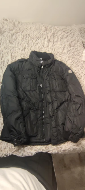 Svart pufferjacka från Moncler - Svart pufferjacka från Moncler med hög krage, flera fickor med knappar och dragkedja framtill. Jackan har Moncler-logga på ärmen och är tillverkad i polyester. Perfekt för kalla dagar och har en snygg, lite loose passform.