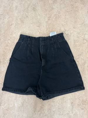 Svarta högmidjade shorts från Zara - Svarta denimshorts från Zara med hög midja och elastisk linning. Modellen har två framfickor och två bakfickor, samt breda benslut med lätt uppvikt kant. Perfekt för en chill och stilren look under varmare dagar.