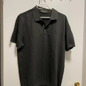 Grön pikétröja från Loro piana  - Säljer en stilren grå pikétröja från Loro piana . Tröjan har en klassisk krage och knappar framtill. Perfekt för en avslappnad men ändå uppklädd look. Oanvänd i 10/10 skick.