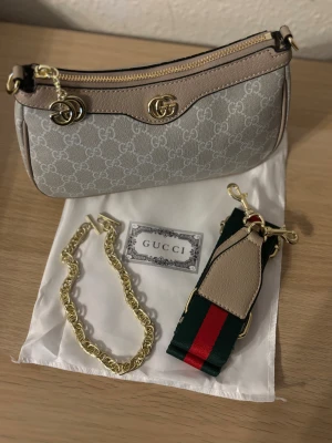 Beige handväska från Gucci - Säljer en snygg handväska från Gucci i beige och grått med klassiskt GG-mönster. Väskan har guldfärgade detaljer, dragkedja och medföljande axelrem i grönt och rött. Materialet är en mix av skinn och textil, perfekt för dig som gillar lyxiga accessoarer.