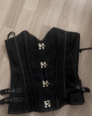 Svart korsett med spännen - Säljer en svart korsett med snygga metallspännen framtill och dekorativa remmar på sidorna. Korsetten har en v-ringad form upptill och är tillverkad i ett mönstrat tyg med detaljer i syntetmaterial. Perfekt för att skapa en edgy look.