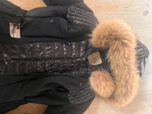 Svart Moncler dunjacka med päls - Säljer en svart Moncler dunjacka med quiltade detaljer och fluffig brun päls på huvan. Jackan har bälte i midjan, dragkedja framtill och långa ärmar. Materialet är dun, polyester och genuin mink-päls. Perfekt julklapp🎅köpt på NK för 16000kr. 9/10 cond