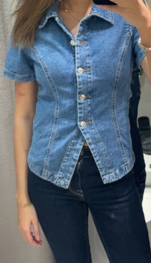 Blå jeanstopp med korta ärmar - Säljer en blå jeansjacka med korta ärmar och snygga sömmar som ger en cool form. Jackan har klassisk krage och knappar framtill. Perfekt för dig som gillar denim och vill ha en jacka som sticker ut lite extra.