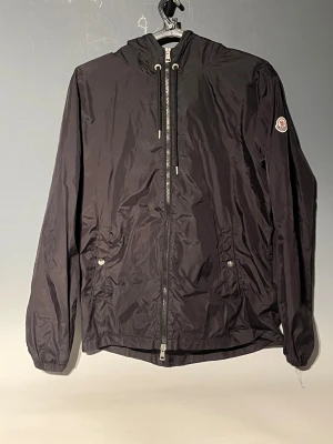 Svart vindjacka från Moncler - Svart vindjacka från Moncler med fungerande nfc och qr. Jackan har Moncler-logga på vänster ärm och snygga detaljer med snörning vid halsen. Materialet är lätt och syntetiskt, perfekt för blåsiga dagar. Röd, vit och blå rand på huvan ger extra stil.