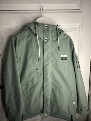 Grön vindjacka från Dope med huva - Snygg ljusgrön vindjacka från Dope med justerbar huva och vita snören. Jackan har dragkedja, flera fickor och coola detaljer som logga på bröstet och ärmen. Perfekt för blåsiga dagar och outdoor vibes.