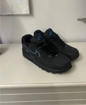 Nike Air Max 90 svart/blå sneakers - Svarta Nike Air Max 90 sneakers med blå detaljer och logga. Skorna har en klassisk siluett med snörning, platt sula och synlig Air Max-dämpning. Ovandelen är i textil och syntet med läderpaneler. Perfekta för dig som gillar streetwear och sportig stil.