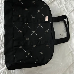 Svart weekenväska från Victoria's Secret - Snygg svart weekendväska från Victoria's Secret med ett diskret mönster av små prickar och logotyper. Väskan har två rejäla handtag och en dragkedja för säker förvaring. Nypris 858 kr