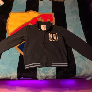 Svart bomberjacka Djurgården - Svart bomberjacka från Djurgården med broderad D-logga på bröstet och '1891' broderat i nacken. Jackan har ribbade muddar med grå och vita ränder, knappar framtill och två fickor. Snygga broderade detaljer på ryggen med text om Djurgården.