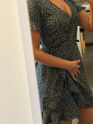 Svart prickig omlottklänning - Säljer en svart kort omlottklänning med vita prickar och volangkant. Klänningen har v-ringning, knyt i midjan och korta ärmar. Perfekt för dig som gillar en lekfull och trendig stil.