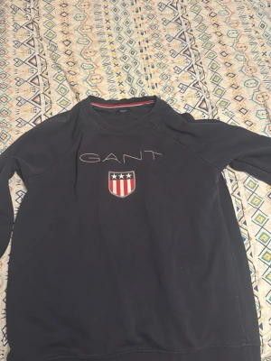 Svart långärmad tröja från GANT - Svart långärmad tröja från GANT med broderad logga och ett snyggt emblem med stjärnor och ränder på bröstet. Tröjan har rund hals och ribbade muddar vid ärmslut och nederkant. Perfekt för dig som gillar klassisk och sportig stil.