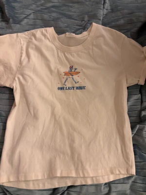 Beige t-shirt med surftryck - Snygg beige t-shirt från Viver med tryck av ett skelett som bär en surfbräda och texten 'One Last Wave' på bröstet. Klassisk rund hals och korta ärmar. Perfekt för dig som gillar chill och surfvibes.