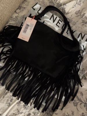 Svart axelväska med fransar från Nelly - Helt ny francis väska från Nelly! Prislapp finns kvar och den är oanvänd. Säljer då den inte föll mig i smaken som jag hade hoppats. Väskan är i en variant av mocka och är en casual men snygg detalj till outfiten. Inuti finns det ett smidigt litet innerfack och väskan sluts med ett magnetknäppe. Sedan kan man stänga remmarna med ett stål spänne ( se bild 5 ) Ord. Pris: 399kr medan jag säljer den för 255kr inklusive frakt. 💖