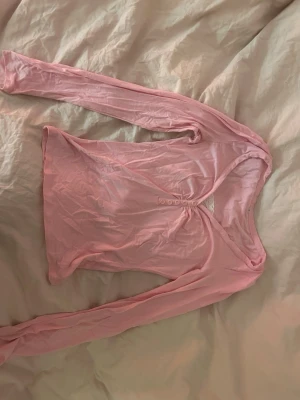 Ljusrosa omlottopp med knappar - Säljer en ljusrosa omlottopp med lång ärm och v-ringning. Toppen har fem dekorativa knappar framtill och en snygg spetsdetalj vid urringningen. Materialet känns mjukt och lätt, troligen bomull eller polyester. Perfekt för dig som gillar romantiska detaljer.