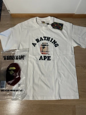 Vit t-shirt från A Bathing Ape M - Säljer en clean vit t-shirt från A Bathing Ape med klassisk logga och rutigt ape-head print på bröstet. T-shirten har rund hals, korta ärmar och tag med ape-loggan på sidan. Materialet är mjuk bomull och medföljer originalförpackning och tag.