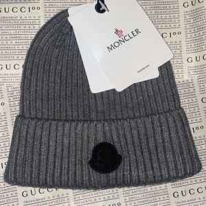 Grå ribbstickad Moncler mössa - Säljer en snygg grå mössa från Moncler med ribbstickad design och svart broderad logga framtill. Mössan är tillverkad i mjukt material som håller dig varm och har en klassisk uppvikt kant. Perfekt för dig som vill ha en stilren accessoar med lyxig känsla.