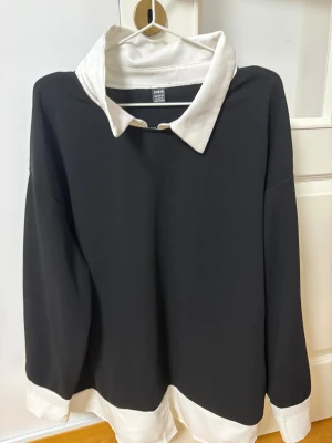 Svart långärmad skjorta från SHEIN - Svart långärmad skjorta från SHEIN i storlek XL med vit krage och vita muddar. Skjortan har en klassisk, loose passform och är tillverkad i syntetmaterial. Perfekt för dig som gillar kontraster och stilrena detaljer.