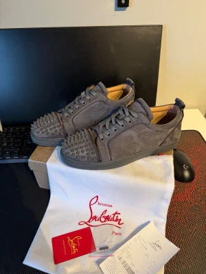 Grå Christian Louboutin sneakers med nitar - Säljer ett par gråa sneakers från Christian Louboutin i mocka med coola nitar på tån och klassisk röd sula. Skorna har snörning och rund tå, och kommer med originalpåse, certifikat och extra nitar. Perfekt för dig som vill sticka ut med lyxig streetstyle. Priset kan diskuteras vid snabbast affär 