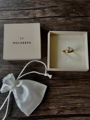 Ring från Mockberg - Som en droppe varm skandinavisk elegans – 18K-guldpläterat stål omfamnar en perfekt droppformad vit cubic zirconia som gnistrar i varje rörelse. Vattentålig design följer med dig genom sommarregn, strandbad eller vardagsduschen utan att tappa sin lyster. Bär den med subtil styrka och känn hur din självsäkerhet strålar varje dag.