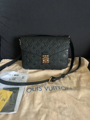 Svart axelväska  - Säljer en svart axelväska med klassiskt monogrammönster i skinn. Väskan har guldfärgade metalldetaljer, justerbar axelrem och ett praktiskt handtag. Insidan är fodrad med brunt tyg med ränder och har flera fack. Äkta skinn och medföljer dustbag. Helt ny 